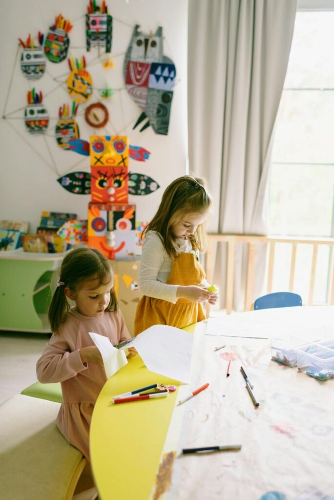 Two young girls crafting in a colorful preschool classroom, surrounded by art supplies and handmade decorations.